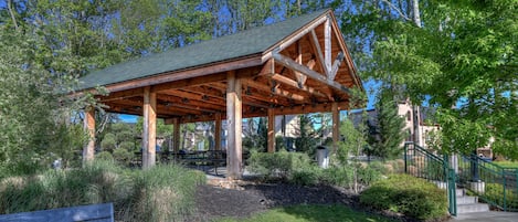 A wooden pavilion with a green roof, surrounded by lush greenery and a bench.