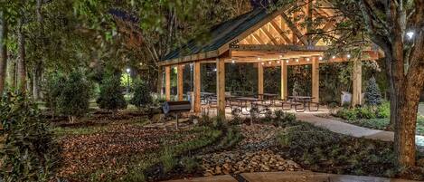 A wooden pavilion with benches and picnic tables, surrounded by a landscaped garden with trees and a stone pathway.