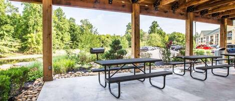 Un área de picnic techada al aire libre con suelo de concreto y vigas de madera, rodeada de vegetación y vista a un estacionamiento.
