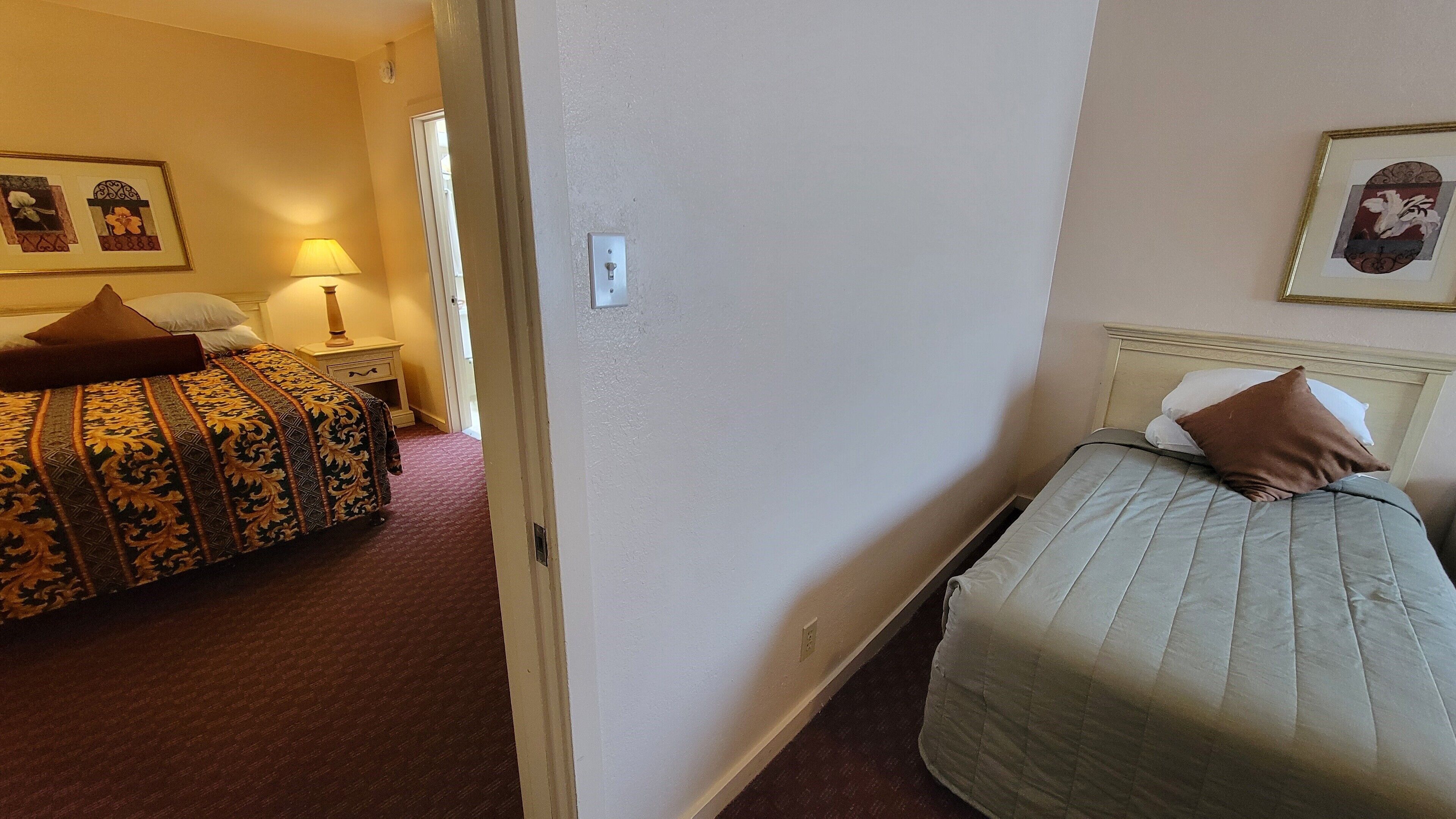 Una habitación de hotel con dos camas, una mesita de noche, una lámpara y una puerta que da a otra estancia.