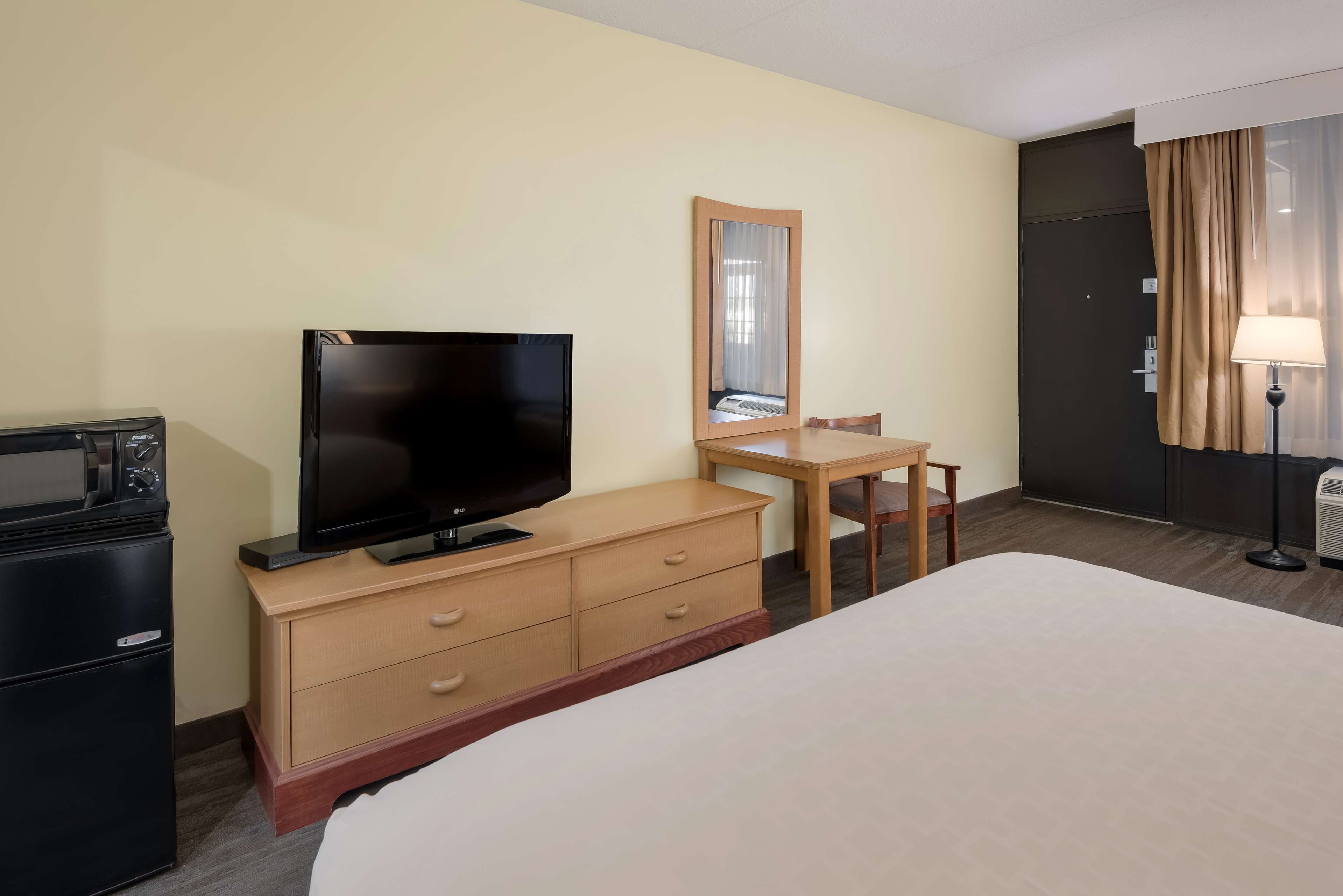 Une chambre d’hôtel comprenant un lit, un téléviseur posé sur une commode en bois, un bureau avec une chaise, un miroir, une lampe et un mini-bar.