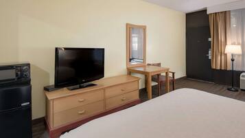 Une chambre d’hôtel comprenant un lit, un téléviseur posé sur une commode en bois, un bureau avec une chaise, un miroir, une lampe et un mini-bar.