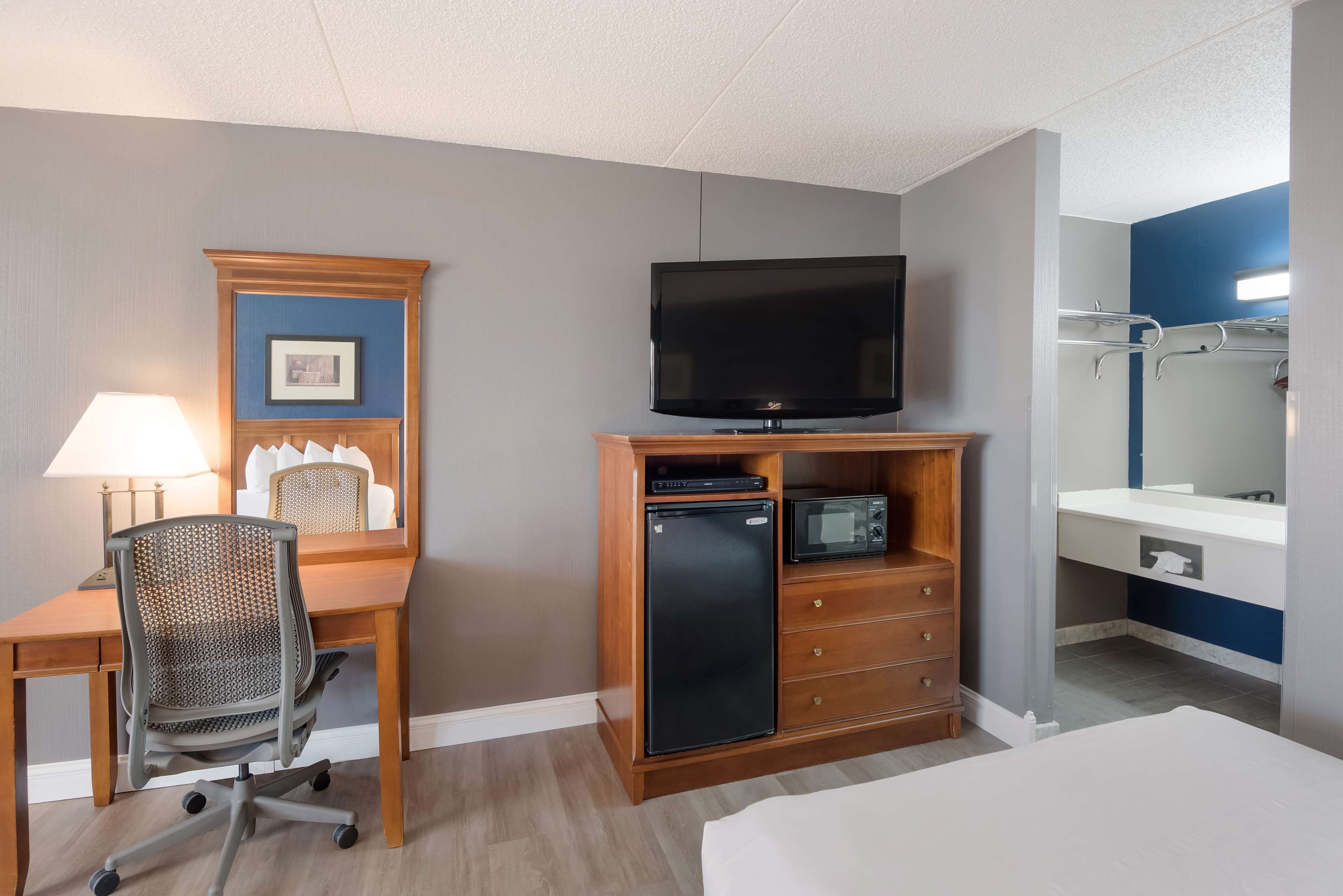 A hotel room with a wooden desk, a television, a small refrigerator, a microwave, a chair, a bed, and a bathroom with a mirror and towel rack.