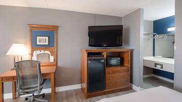 Habitación de hotel con escritorio de madera, televisor, refrigerador pequeño, microondas, silla, cama y baño con espejo y perchero para toallas.
