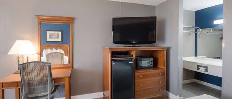A hotel room with a wooden desk, a television, a small refrigerator, a microwave, a chair, a bed, and a bathroom with a mirror and towel rack.