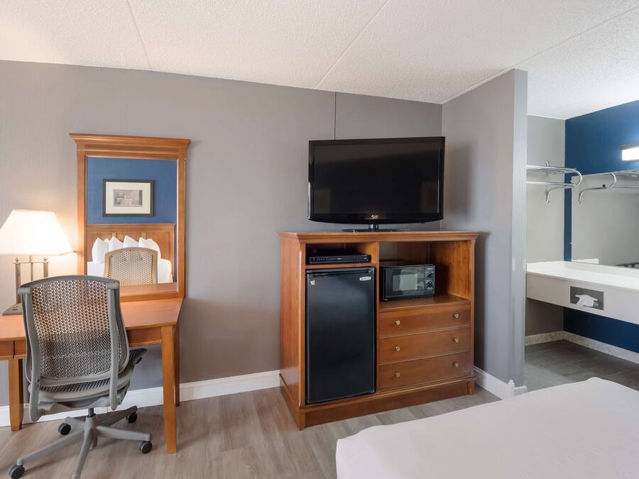 Habitación de hotel con escritorio de madera, televisor, refrigerador pequeño, microondas, silla, cama y baño con espejo y perchero para toallas.