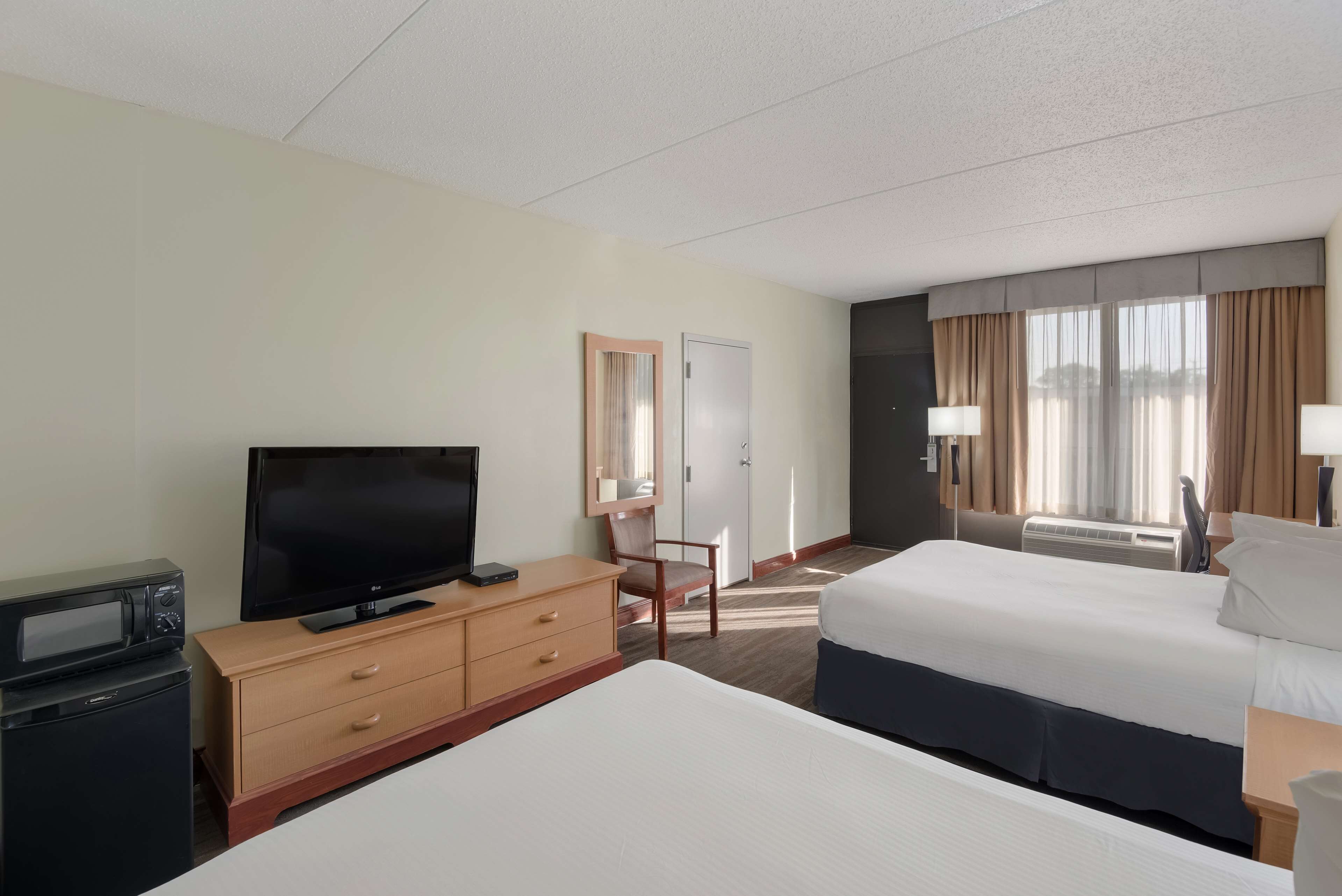 A hotel room with a bed, a TV on a wooden dresser, a desk with a lamp, and a window with curtains.