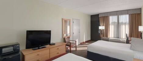 A hotel room with a bed, a TV on a wooden dresser, a desk with a lamp, and a window with curtains.