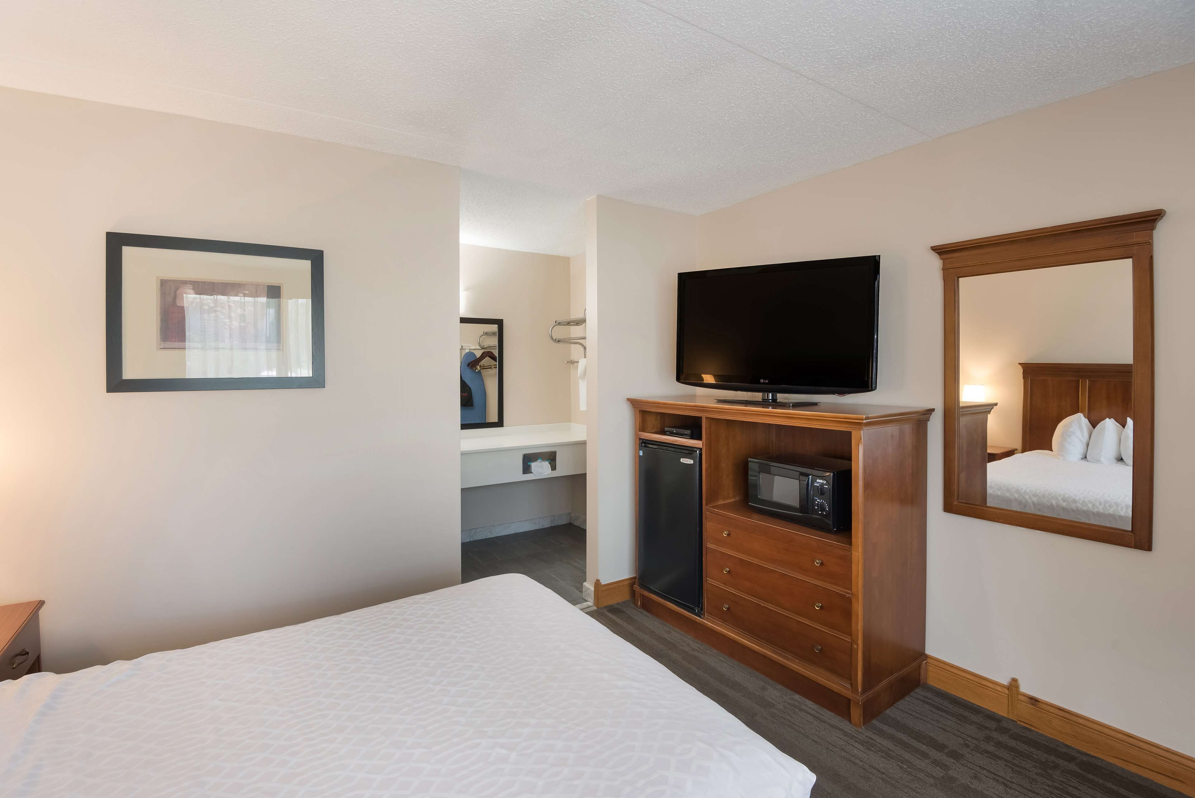 A hotel room with a bed, a television, a microwave, a small refrigerator, a wooden dresser, a mirror, and a framed picture on the wall.
