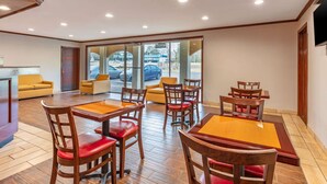Un hall de hotel con piso de madera, un área central de comedor con mesas y sillas, y un gran ventanal que da al exterior.