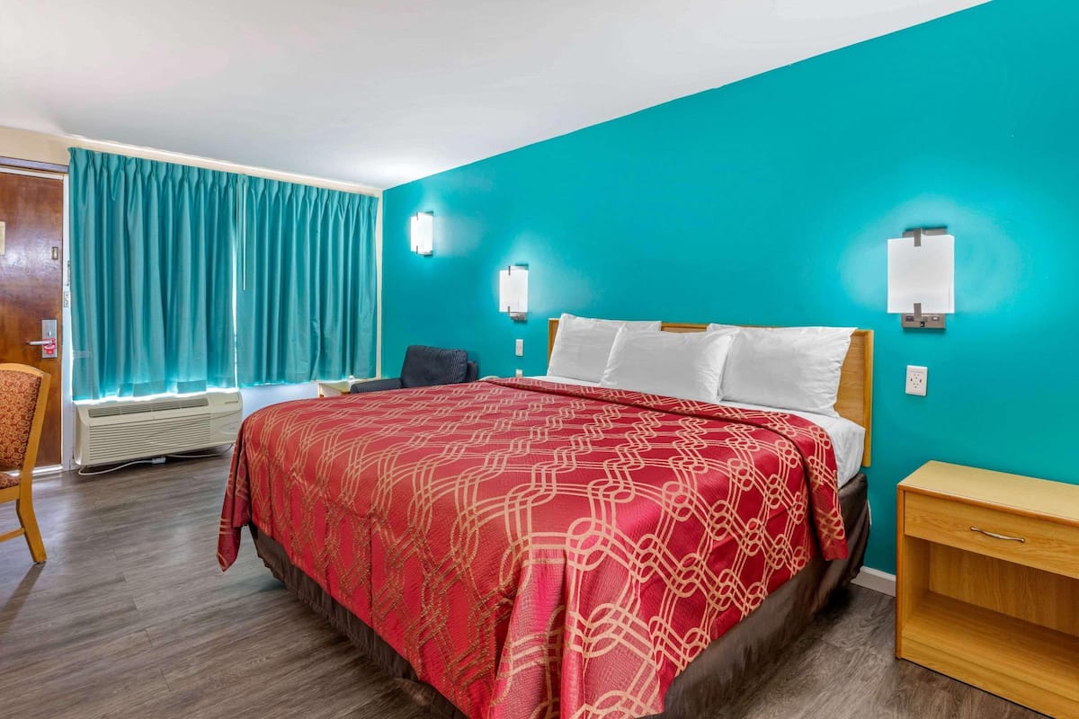 A hotel room with a teal wall, a wooden floor, a bed with a red patterned bedspread, a wooden nightstand, and a chair.