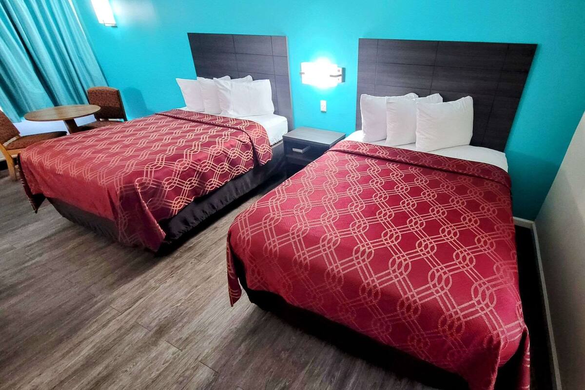 A hotel room with two beds, each with a red patterned bedspread and white pillows. There is a small table and a chair in the corner, and a wall-mounted light fixture above the beds.