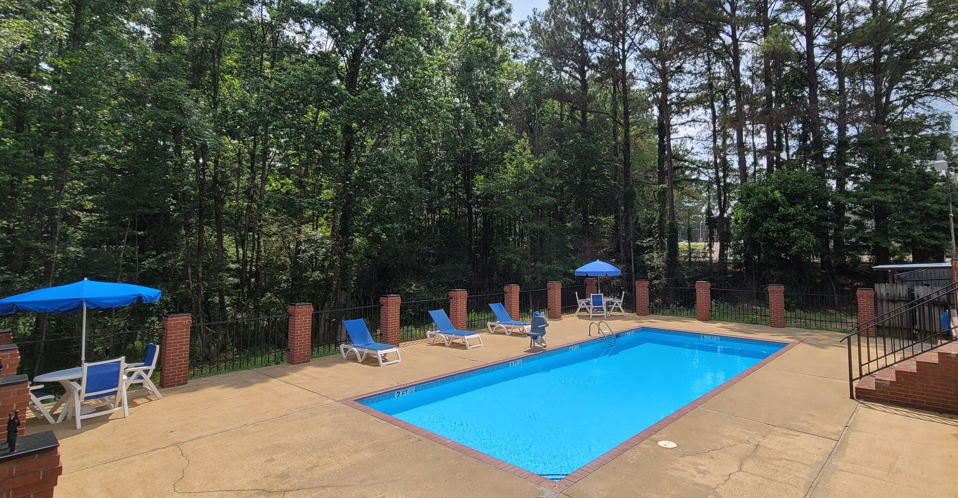A rectangular swimming pool with a blue border, surrounded by a brick patio. There are lounge chairs and umbrellas, and the area is enclosed by a fence with trees in the background.