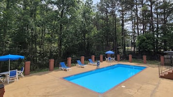 A rectangular swimming pool with a blue border, surrounded by a brick patio. There are lounge chairs and umbrellas, and the area is enclosed by a fence with trees in the background.