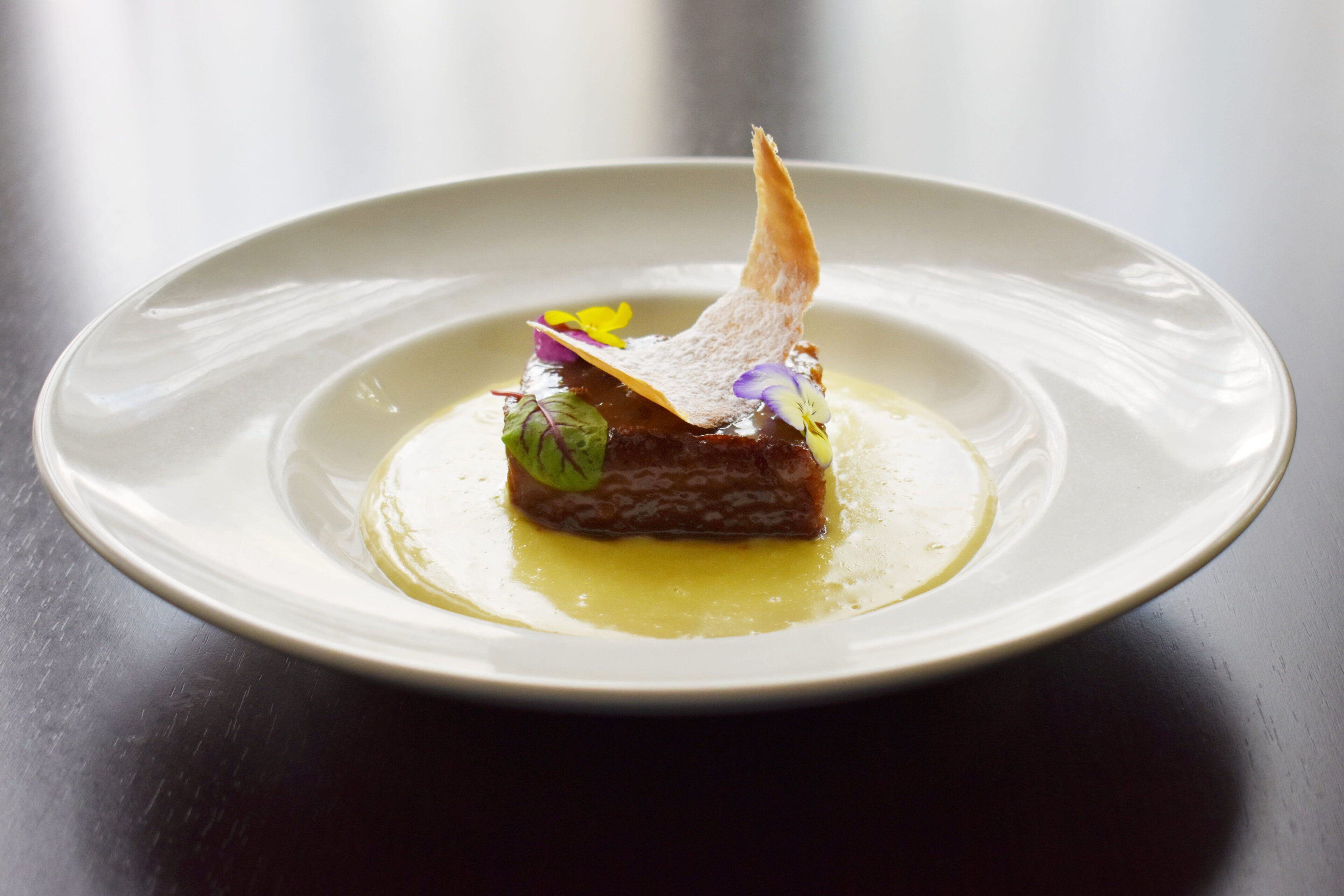 A white plate with a single piece of food garnished with a leaf and a flower, placed on a dark surface.