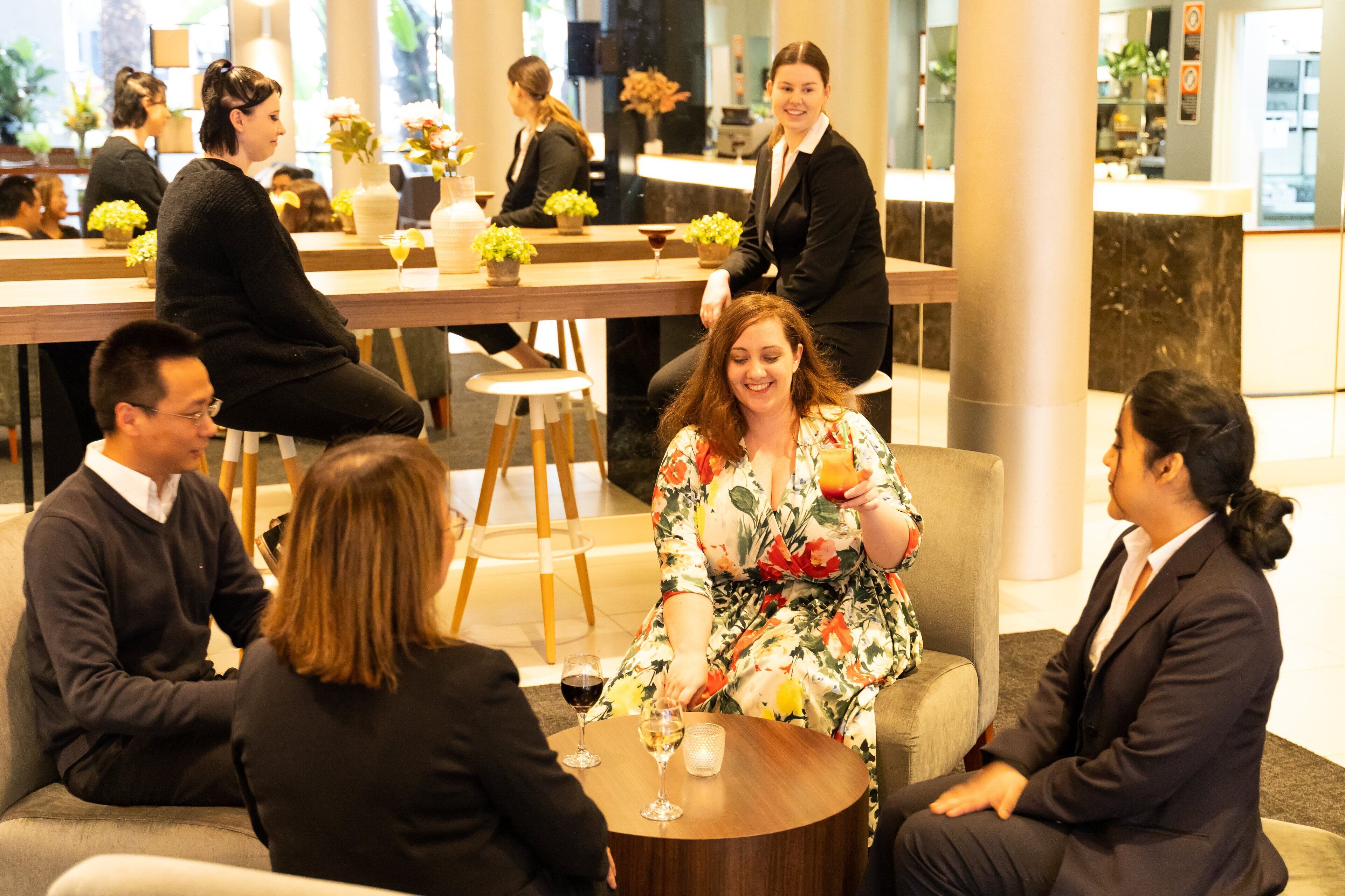 A group of people seated around a round table in a well-lit indoor space with a modern design, featuring a bar area in the background.