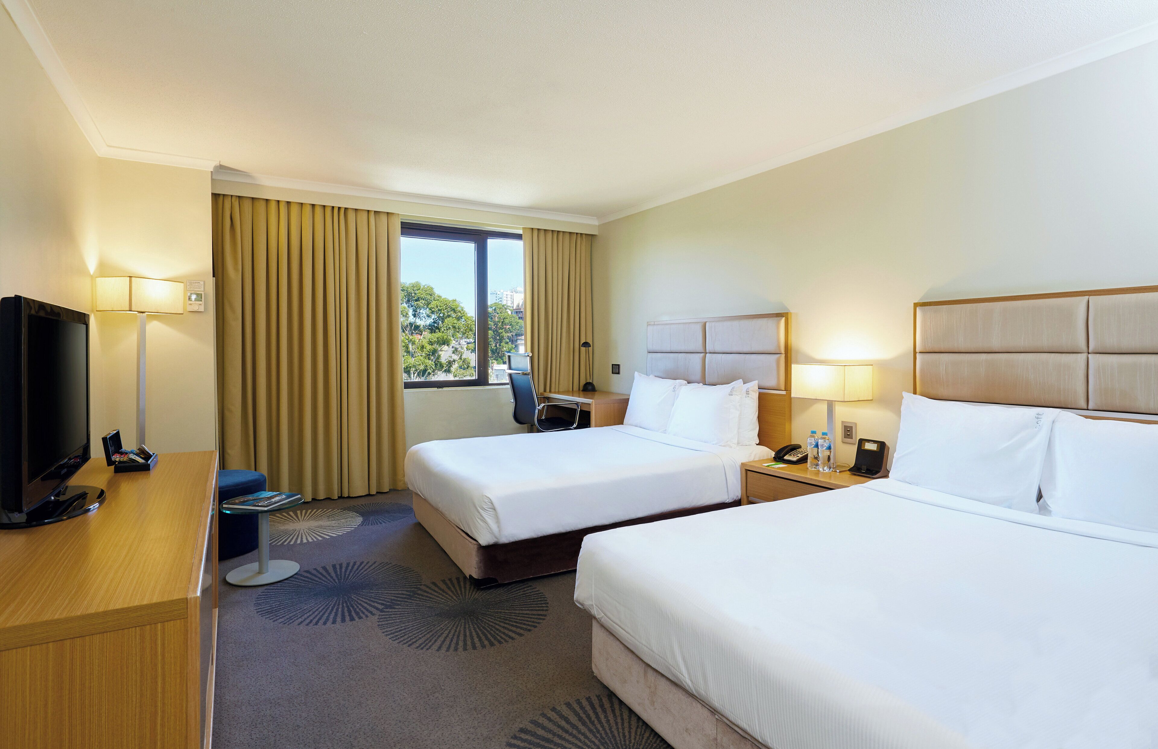 A hotel room with two beds, a desk with a TV, and a window with curtains.