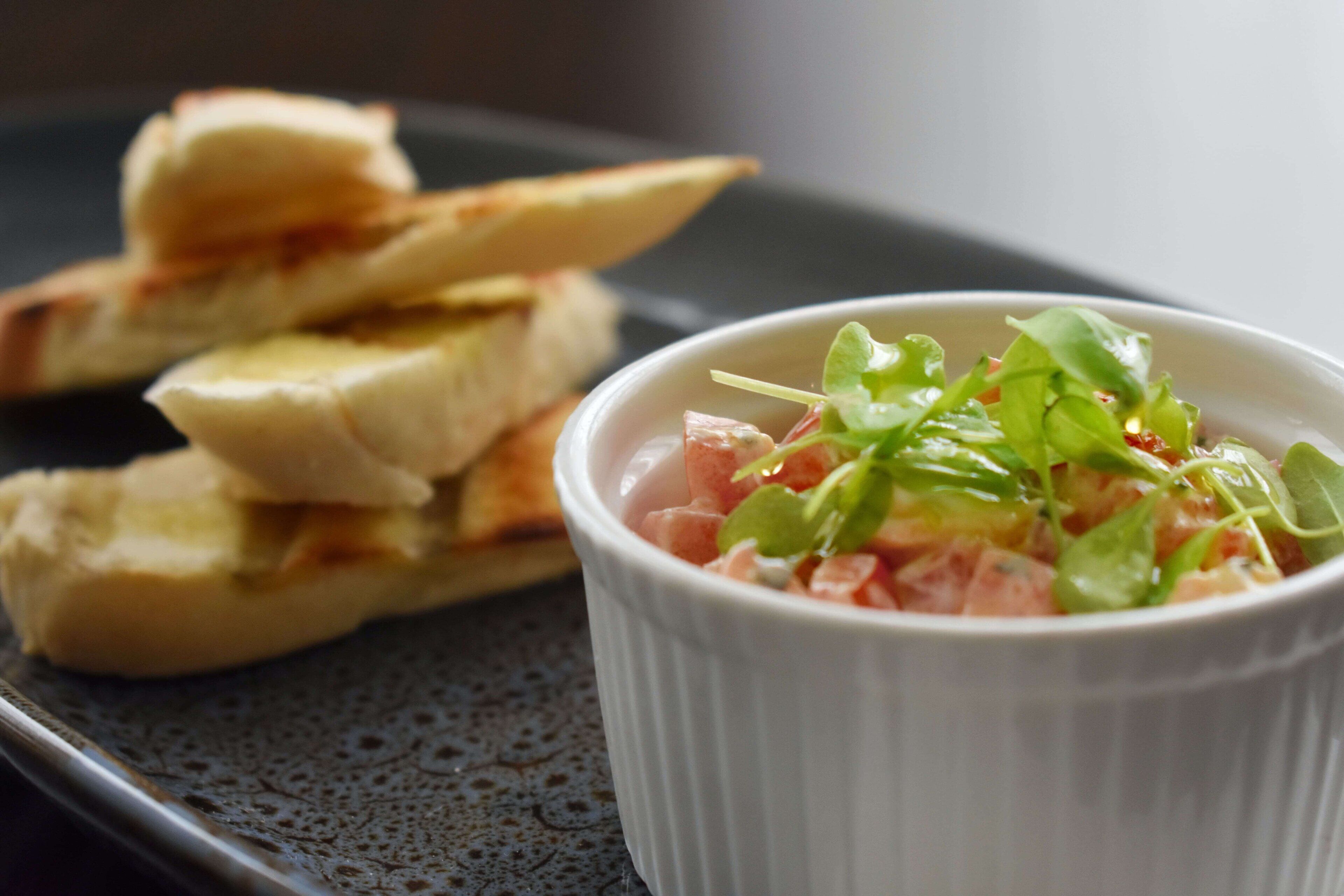 A white bowl of salad with greens and tomatoes on a dark plate, accompanied by a stack of bread.