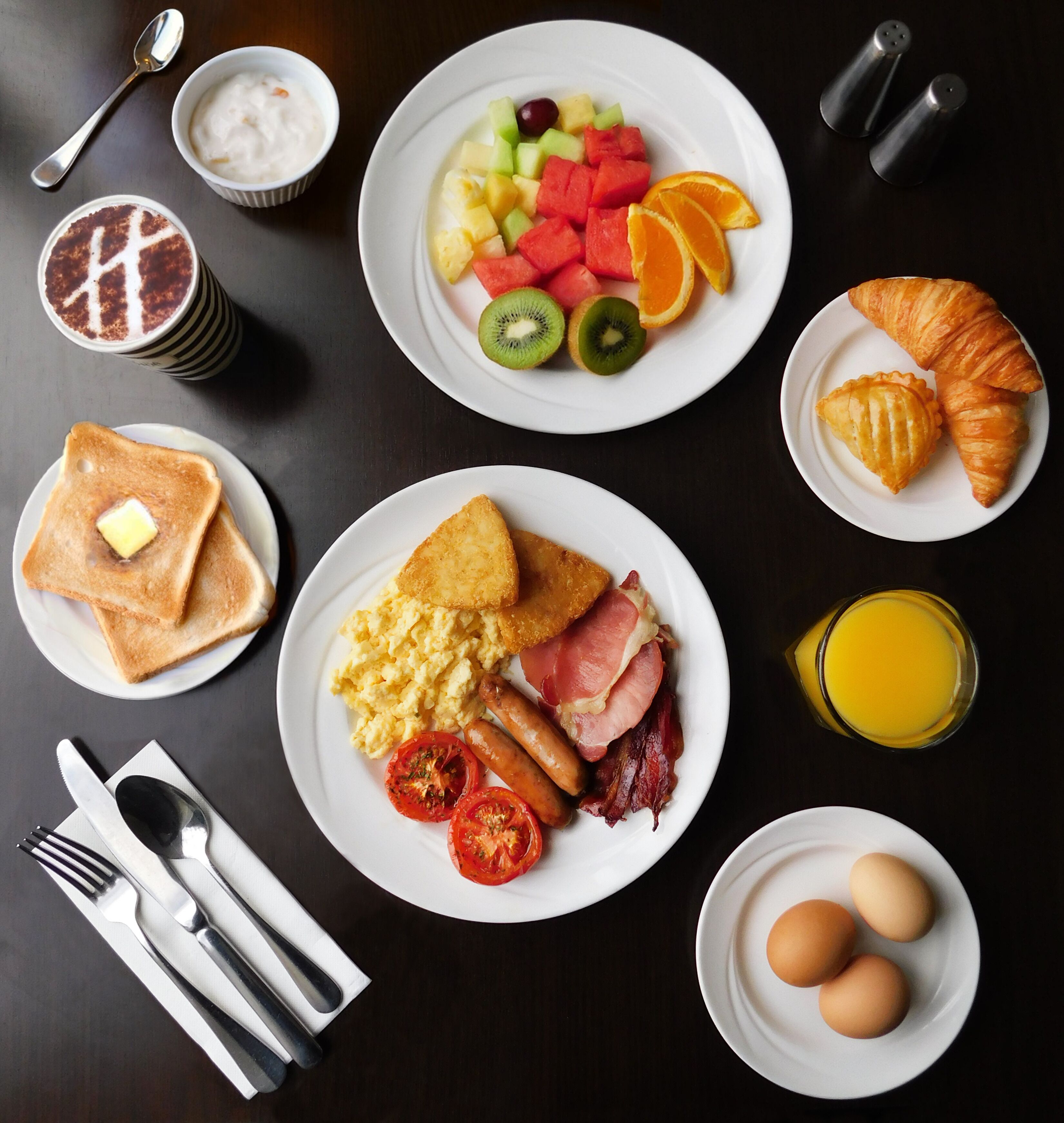 A breakfast spread with toast, scrambled eggs, bacon, tomatoes, sausages, croissants, fruit, and juice.