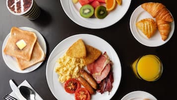 A breakfast spread with toast, scrambled eggs, bacon, tomatoes, sausages, croissants, fruit, and juice.