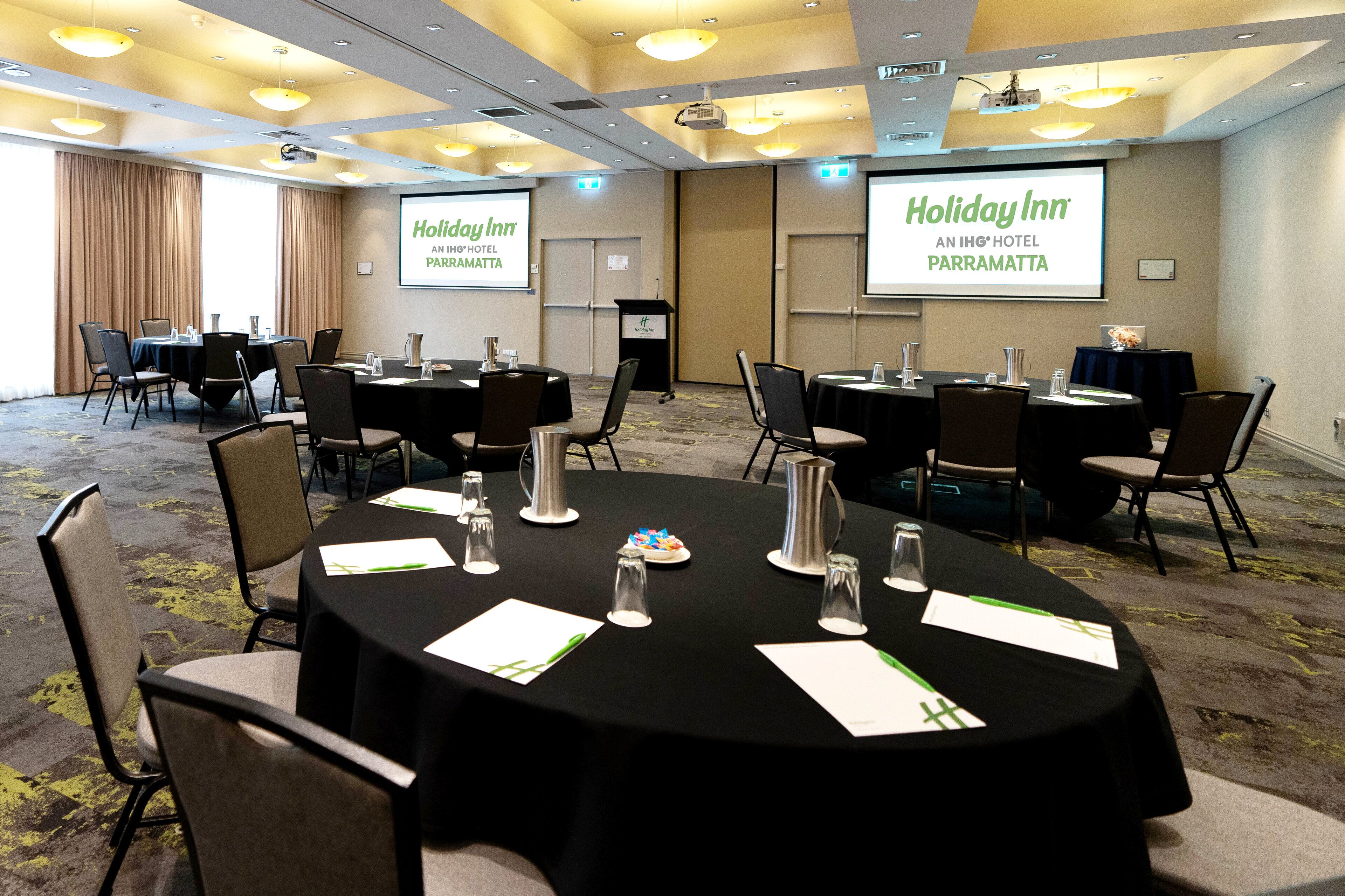 Une salle de conférence équipée de plusieurs tables, de chaises et d’un écran de projection sur lequel est affiché le logo « Holiday Inn ».