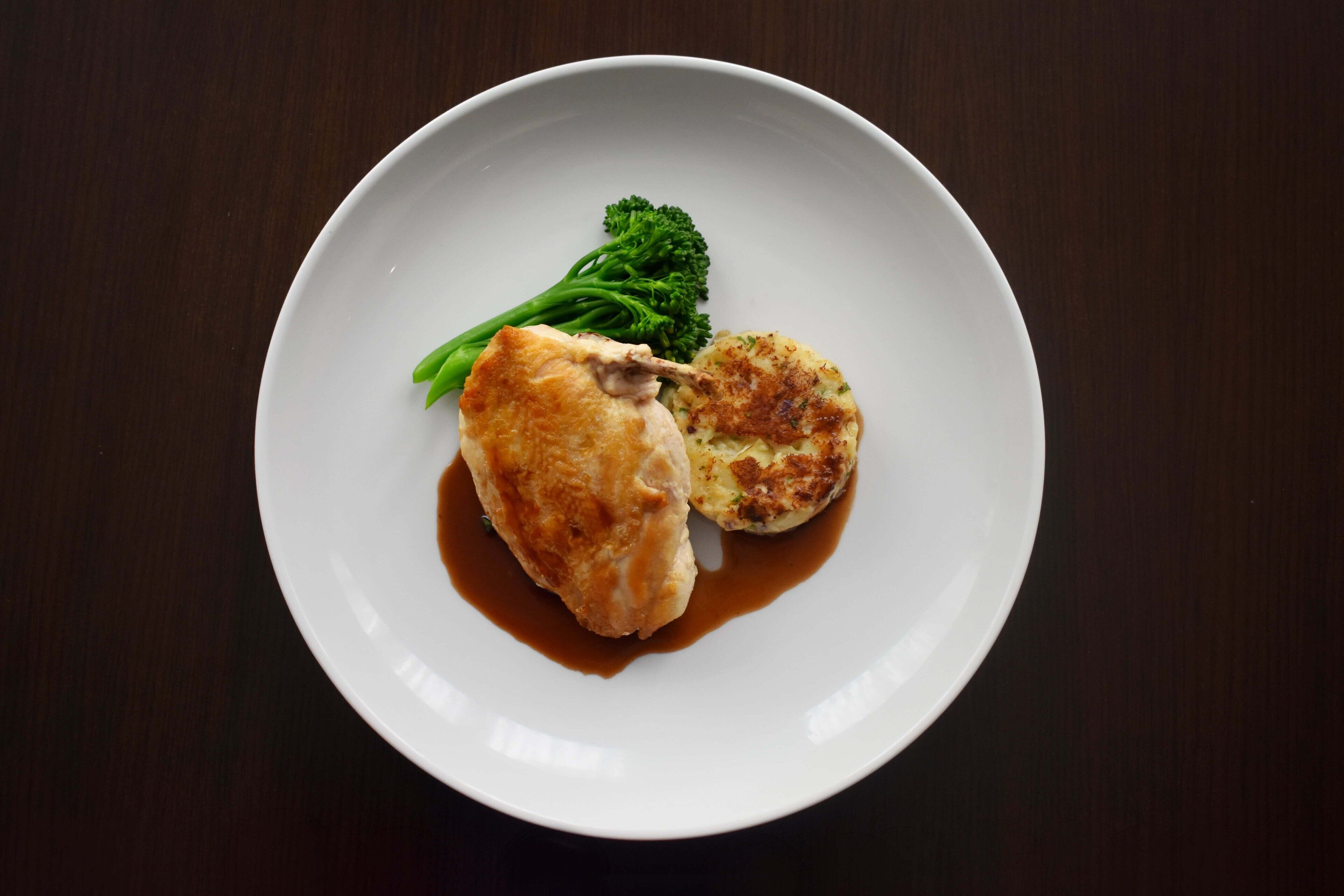 A white plate with a piece of chicken, a round potato, and a small portion of broccoli on a dark wooden table.