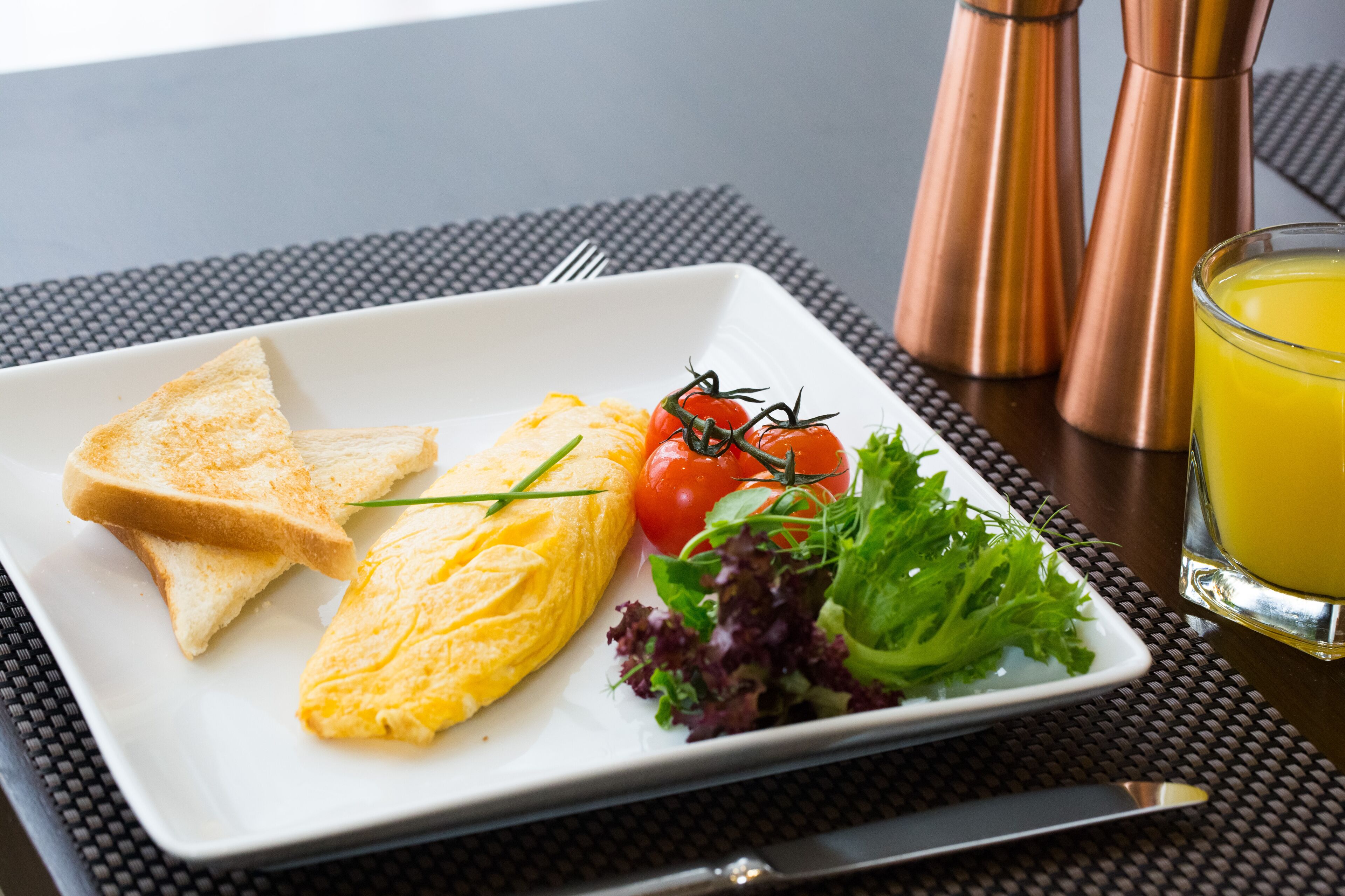 A breakfast plate with toast, scrambled eggs, cherry tomatoes, and greens, accompanied by a glass of orange juice and a pepper grinder.