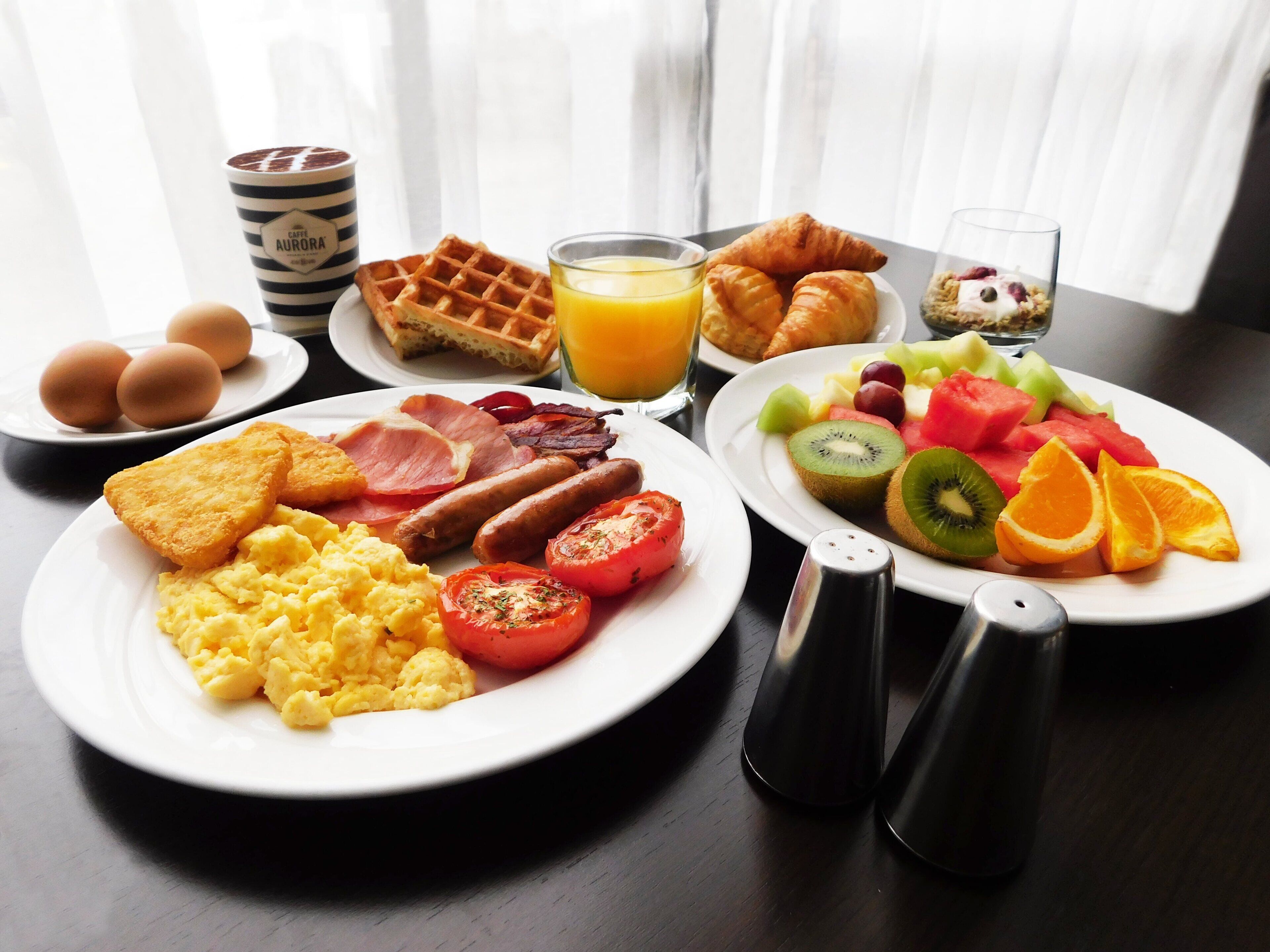 A breakfast table with plates of eggs, bacon, sausages, toast, scrambled eggs, tomatoes, and a variety of fruits including kiwi, watermelon, and oranges. There are also waffles, croissants, and a glass of orange juice.