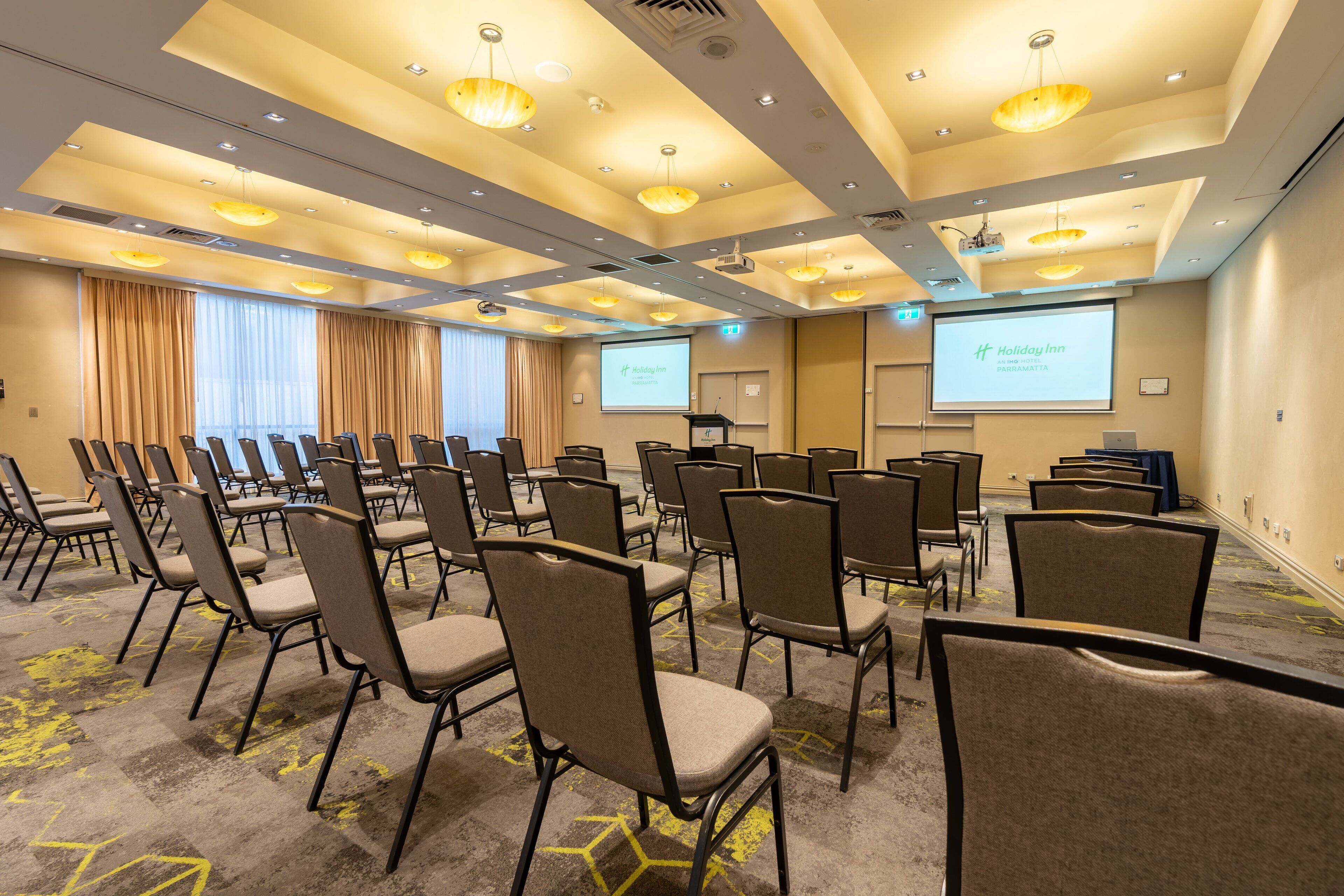 Une salle de conférence avec des rangées de chaises, un écran de projection affichant le nom « Holiday Inn Parramatta », et un tapis orné de motifs géométriques.