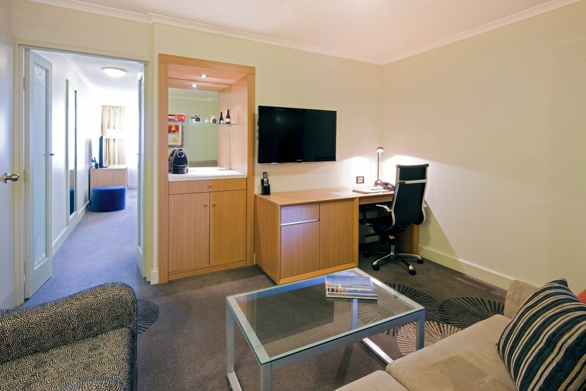 A compact living space with a kitchenette, a flat-screen TV, a desk with a chair, and a sofa.