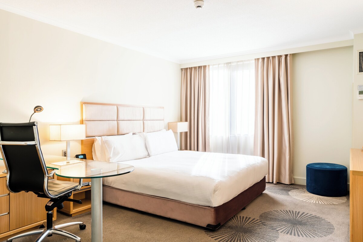 A hotel room with a bed, a desk, a chair, and a lamp.