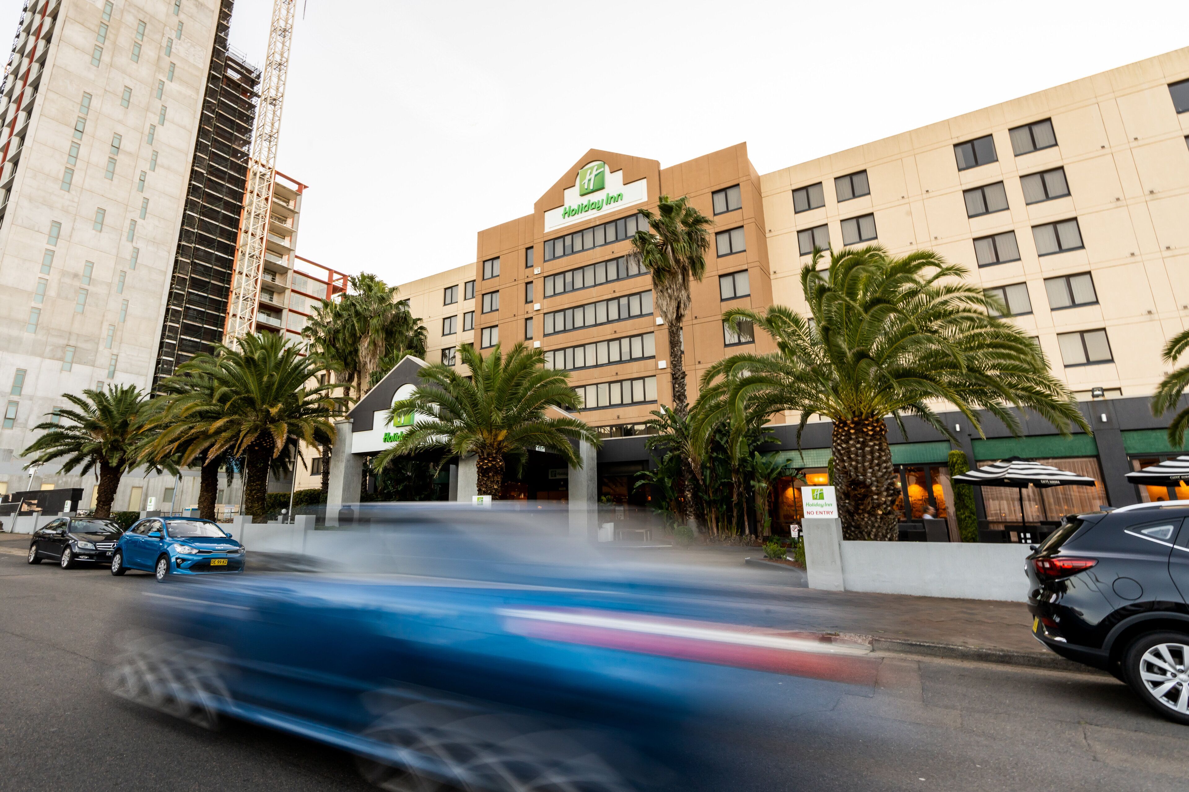 Un hotel Holiday Inn amb un disseny modern, palmeres i un carrer animat amb cotxes.