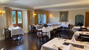 Une salle à manger élégante, avec plusieurs tables dressées pour le service, de grandes fenêtres ornées de rideaux et des luminaires fixés au mur.