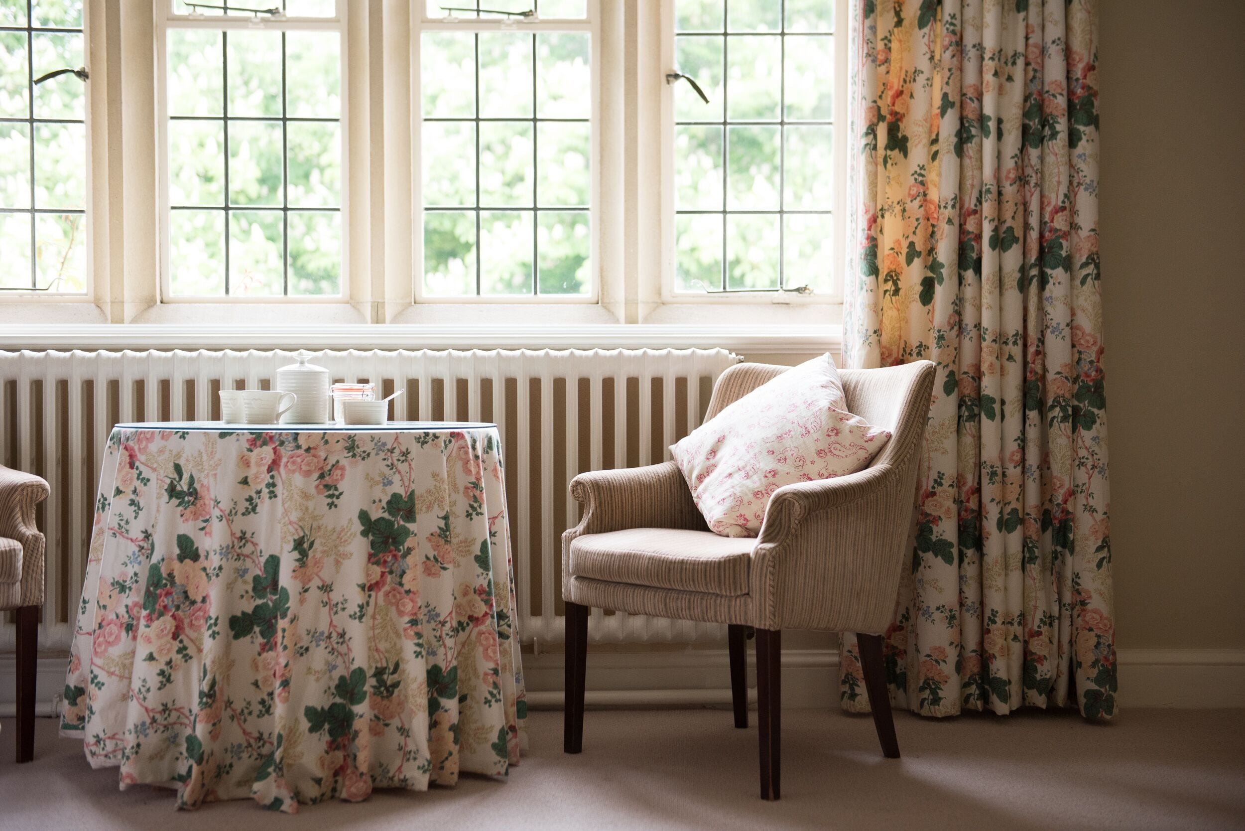 A room with a table covered by a floral tablecloth, a chair with a floral cushion, and a window with a view of greenery.
