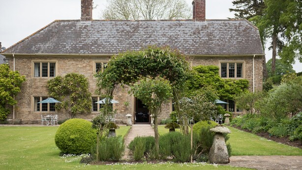 Un bâtiment en pierre à l’architecture traditionnelle, doté d’une entrée centrale avec un passage couvert, entouré de jardins bien entretenus et d’espaces aménagés pour se détendre en plein air.