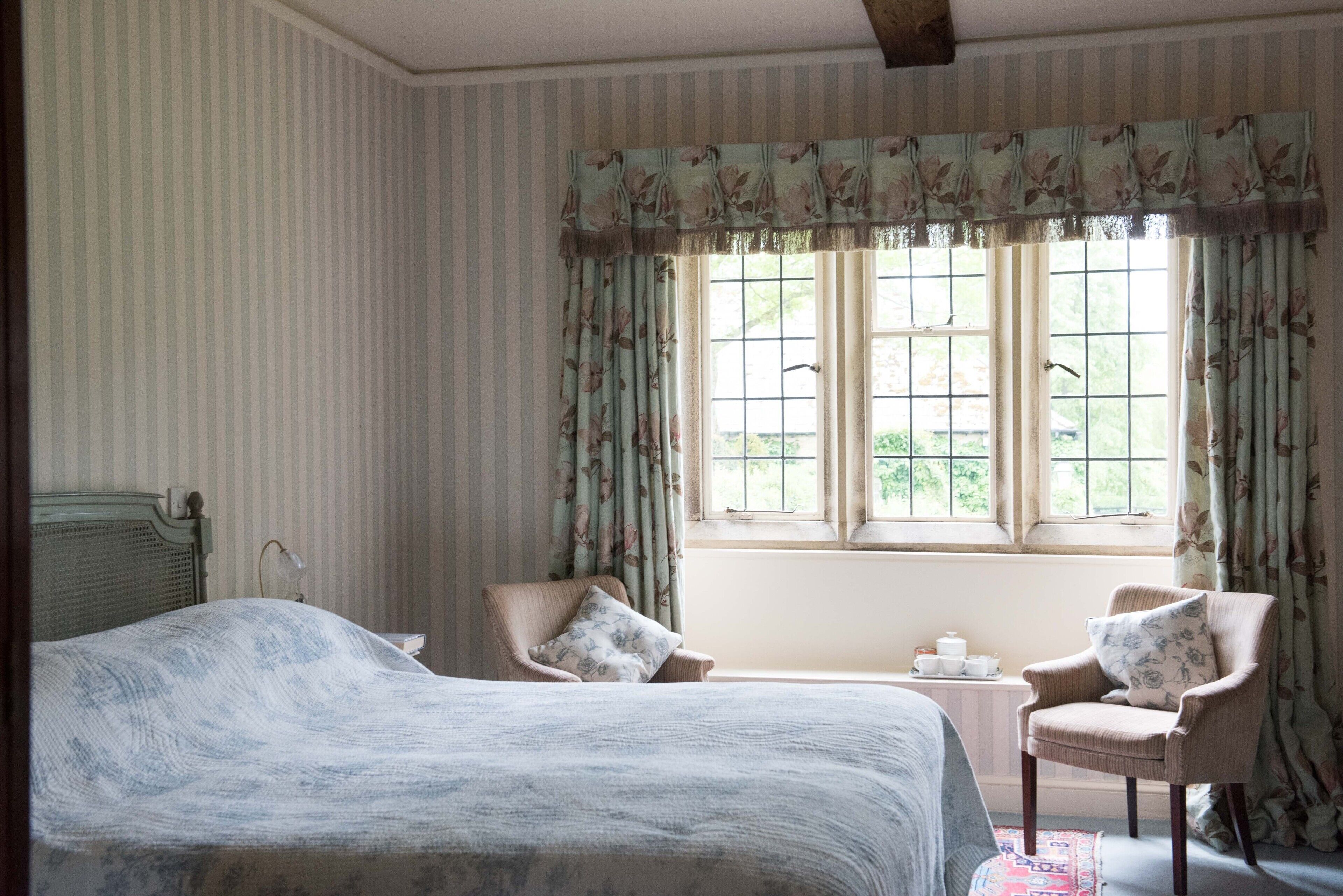 Un dormitorio con una cama grande, dos sillas, una mesita con tetera y una ventana con cortinas.