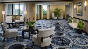 Een hotellobby met een kamerbreed tapijt, zithoek met stoelen en een bank, een klein tafeltje met een plant en een groot raam met gordijnen.