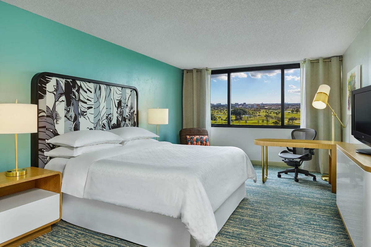 Habitación de hotel con una cama grande, un escritorio con una silla, televisión y ventana con vista a un parque y edificios.