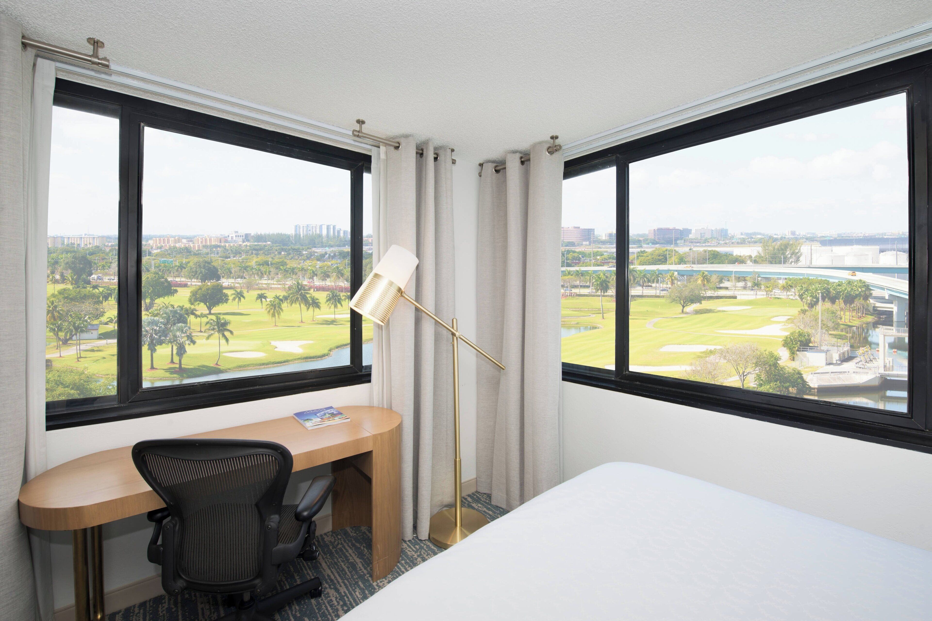 A hotel room with a desk, chair, and a large window offering a view of a golf course and cityscape.