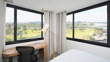 Quarto de hotel com escrivaninha, cadeira e uma grande janela com vista para um campo de golfe e para a paisagem urbana.