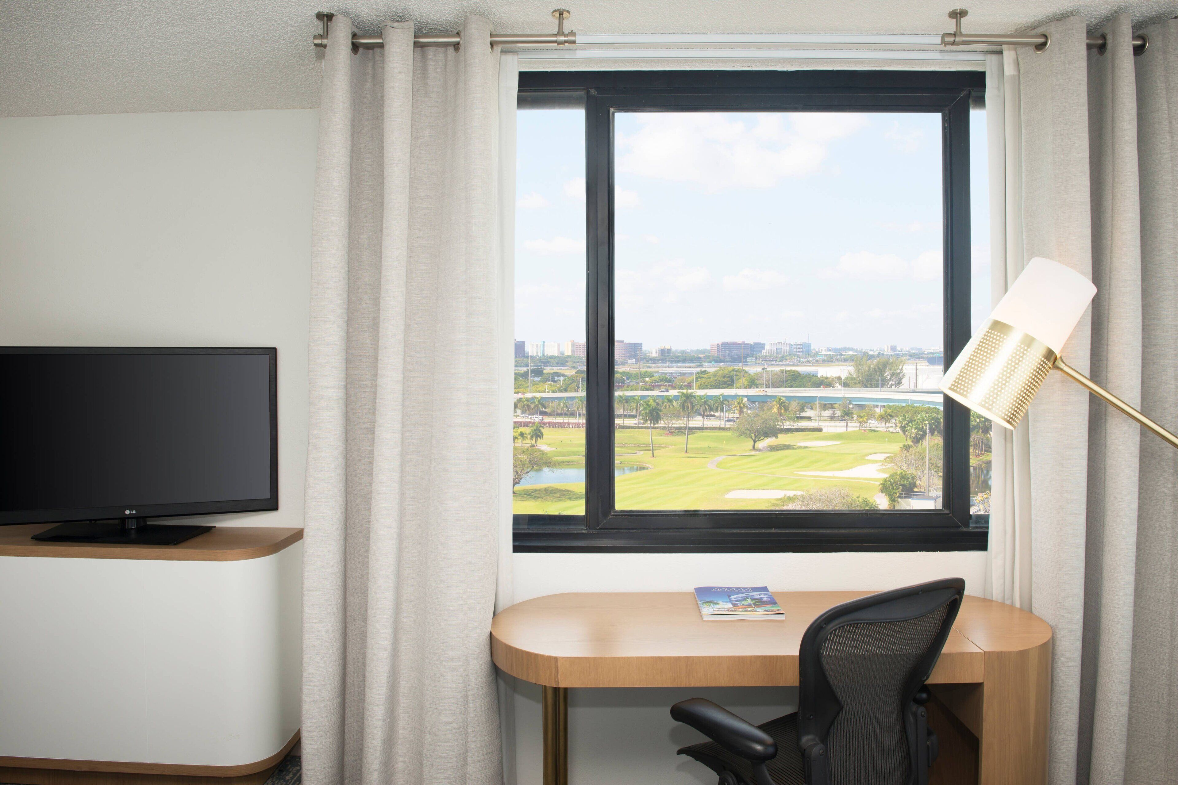 A hotel room with a TV on a stand, a desk with a chair, a lamp, and a window with a view of a golf course and cityscape.