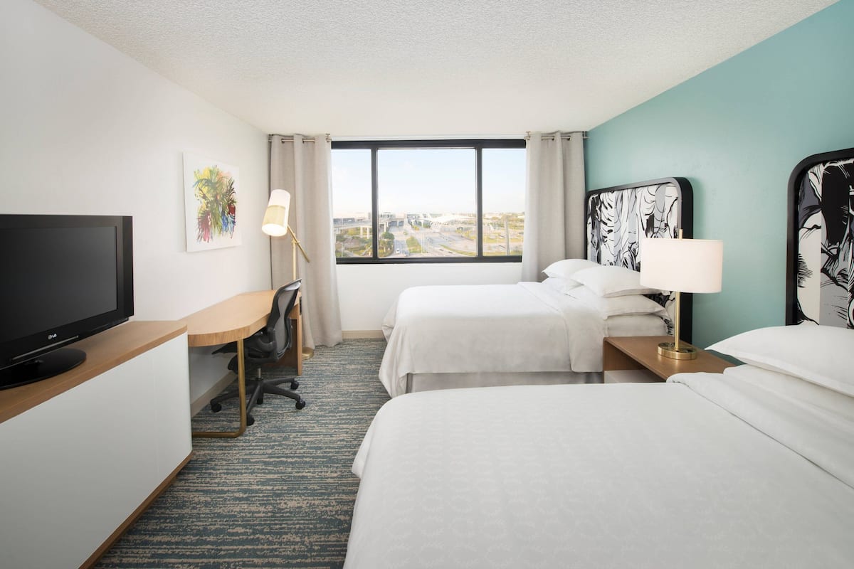 Habitación de hotel con dos camas, un escritorio con televisor, una silla, ventana con vista a la ciudad y obras de arte en las paredes.
