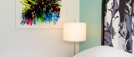 A hotel room with a bed, a nightstand with a lamp, and a colorful abstract painting on the wall.