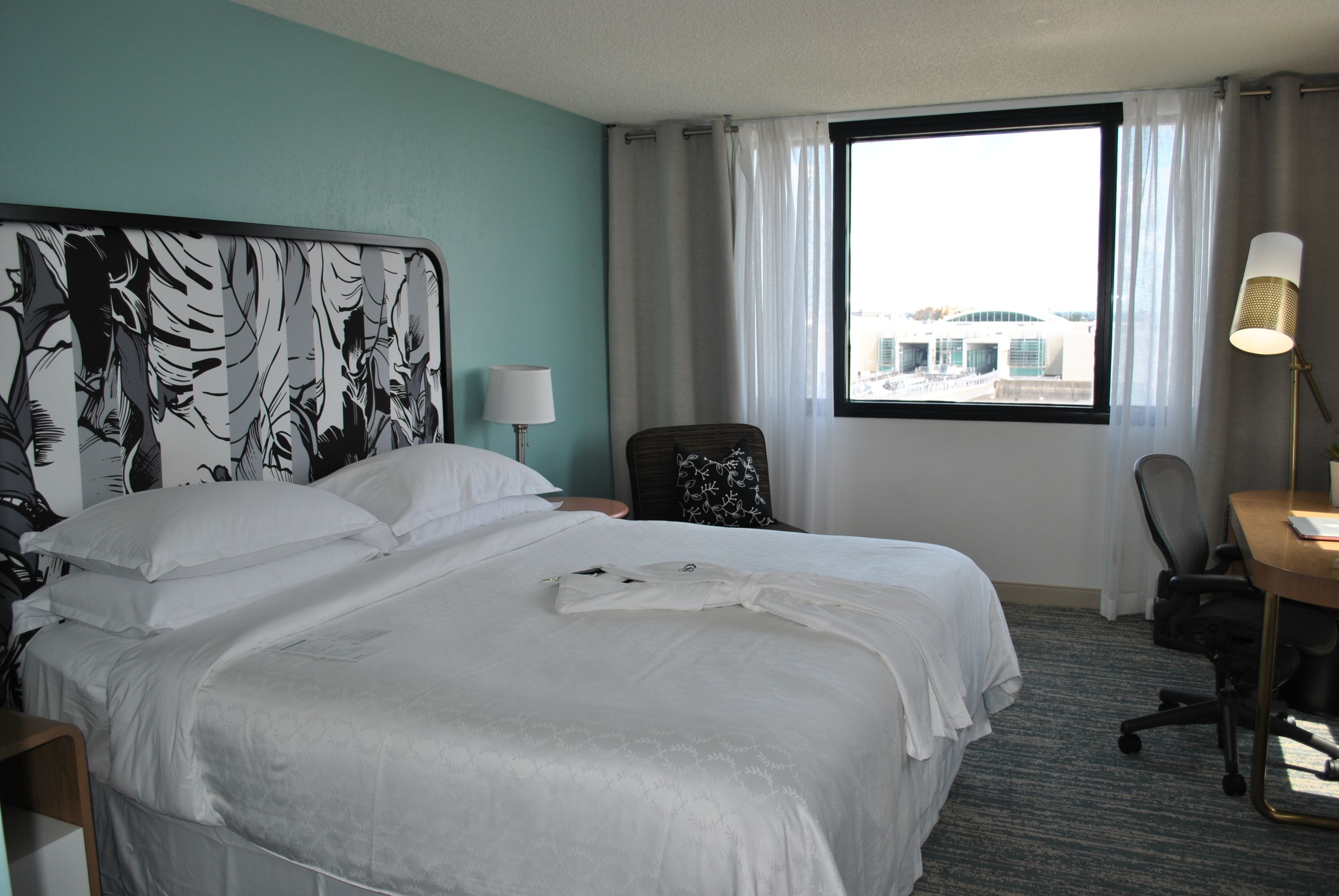 A hotel room with a large bed, a desk, a chair, a lamp, and a window with a view of buildings.