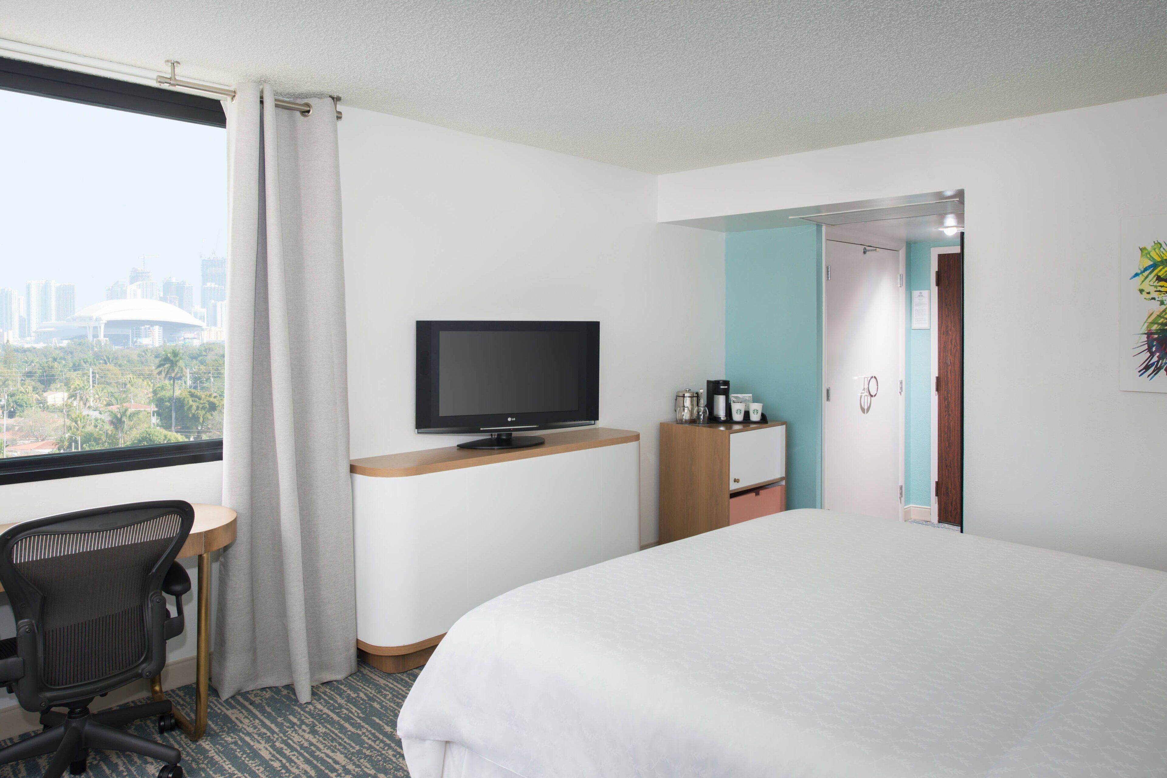A hotel room with a bed, a desk with a computer, a TV, and a view of the city.