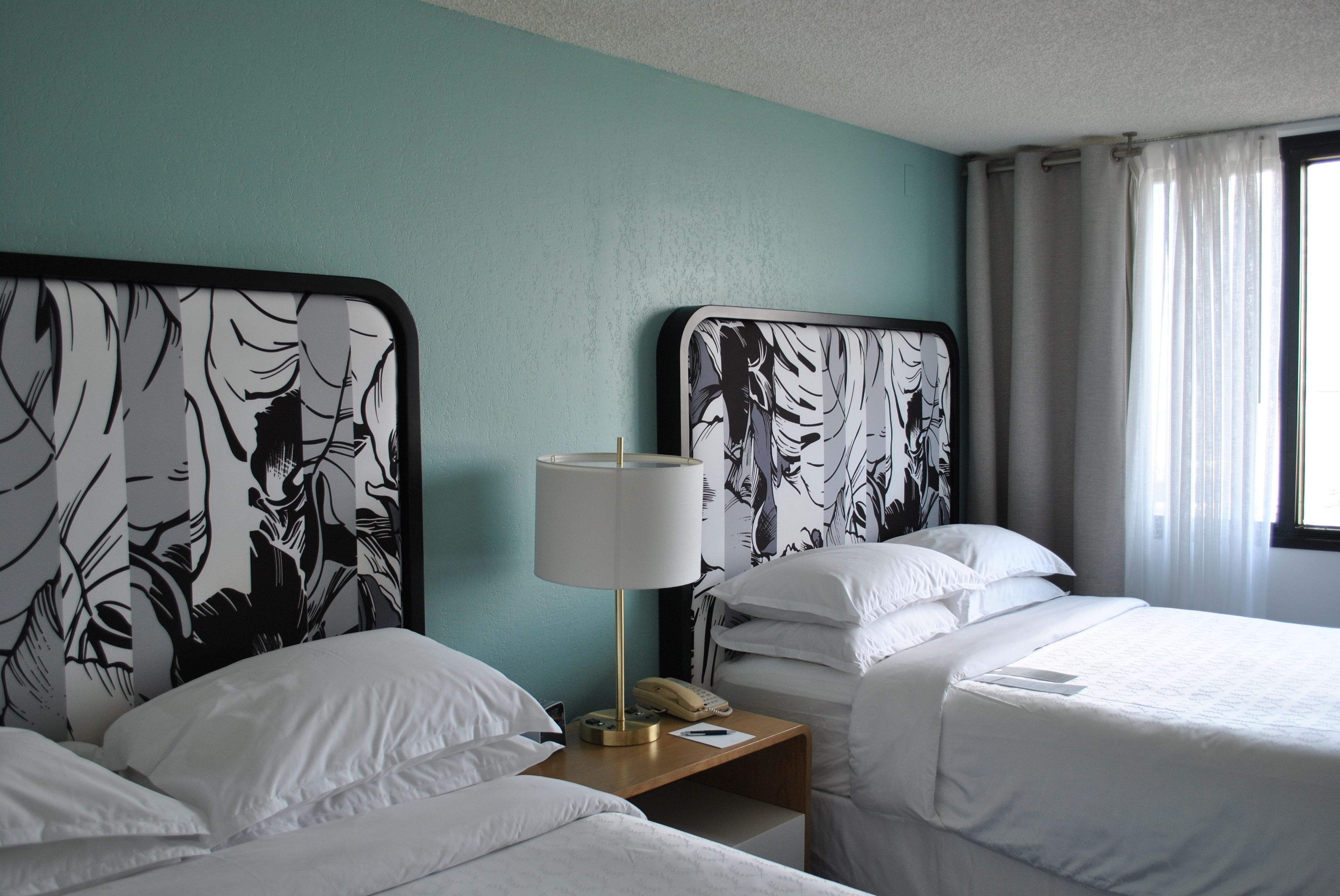 A hotel room with two beds, a nightstand with a lamp, and a window with curtains.