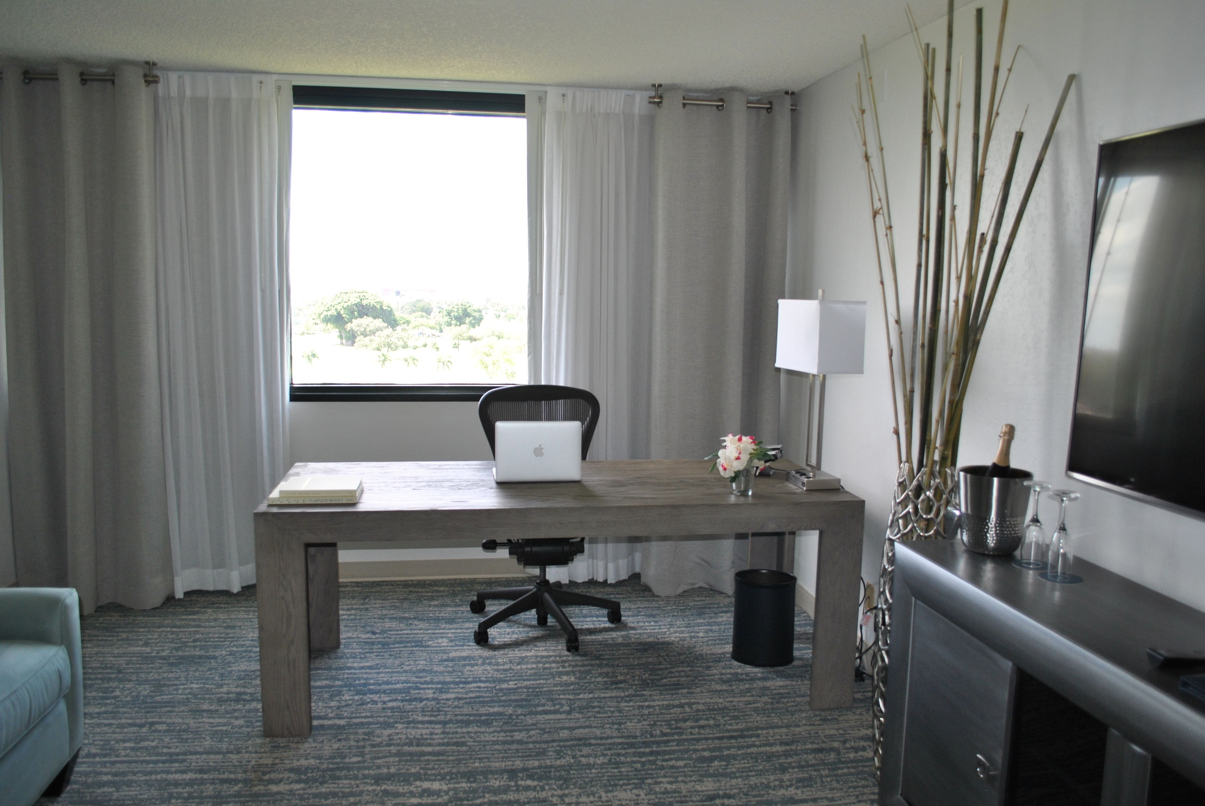 A hotel room with a wooden desk, a laptop, a chair, a TV, a window with curtains, and a vase with flowers.