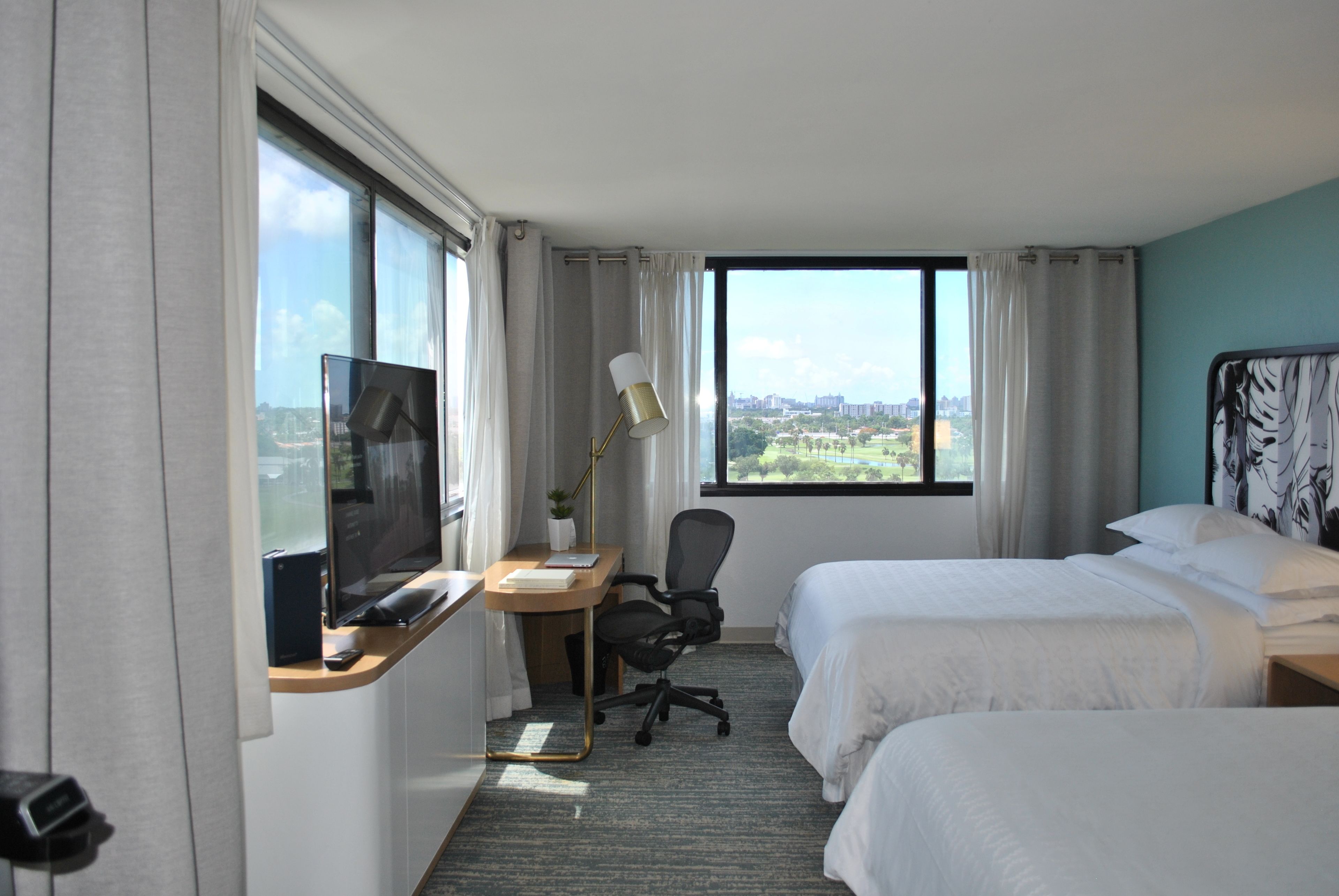 Une chambre d’hôtel avec deux lits, un bureau équipé d’un ordinateur et d’une lampe, une télévision et une vue sur le paysage urbain.