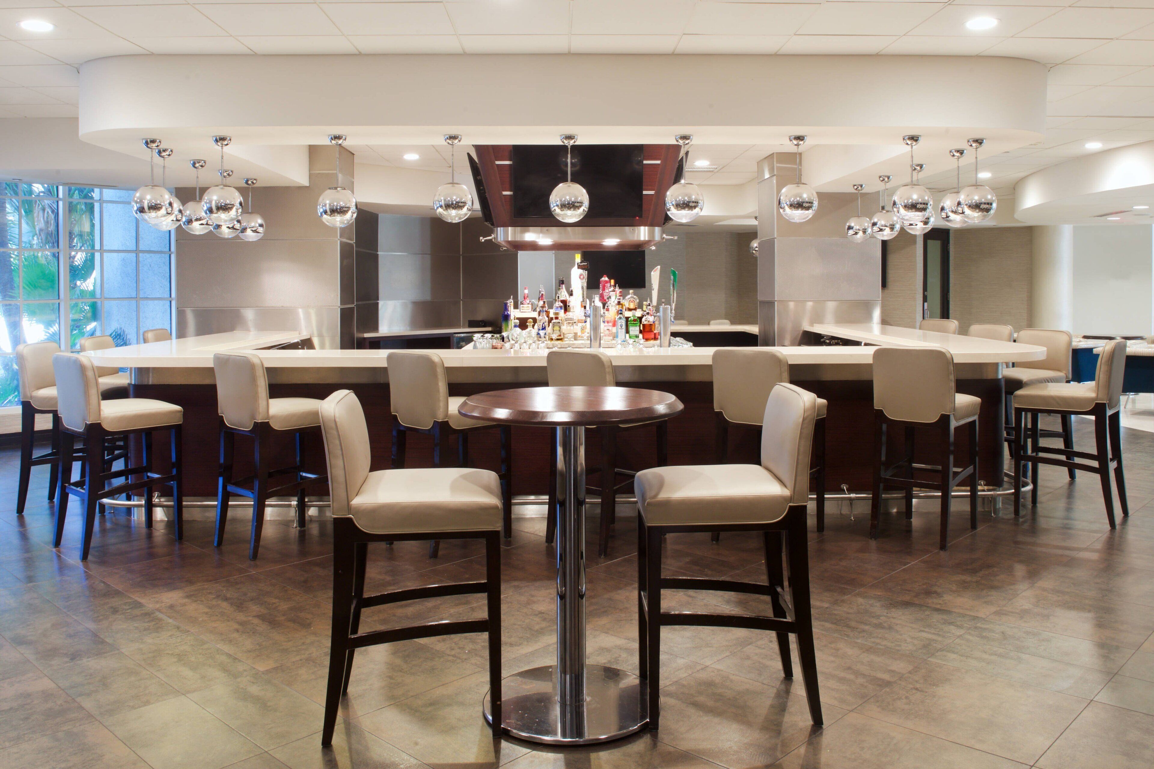 A modern bar area with a central round table surrounded by bar stools, a long counter with a variety of bottles, and a large mirror above the counter.