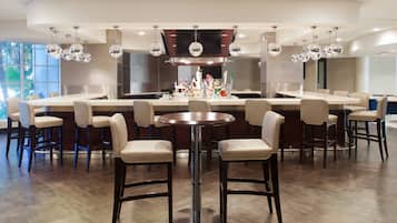 Uma área de bar moderna com uma mesa redonda central rodeada por banquetas, um balcão comprido com uma variedade de garrafas e um espelho grande acima do balcão.